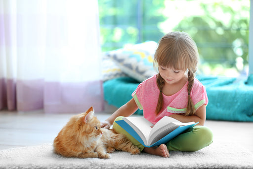 a little girl sitting on carpet, reading her book and stroking her cat