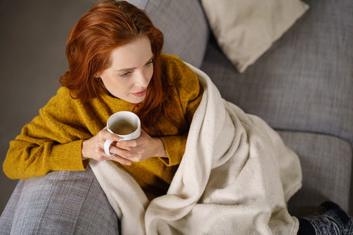 a woman sitting on her couch, drinking tea and wrapped in a warm blanket