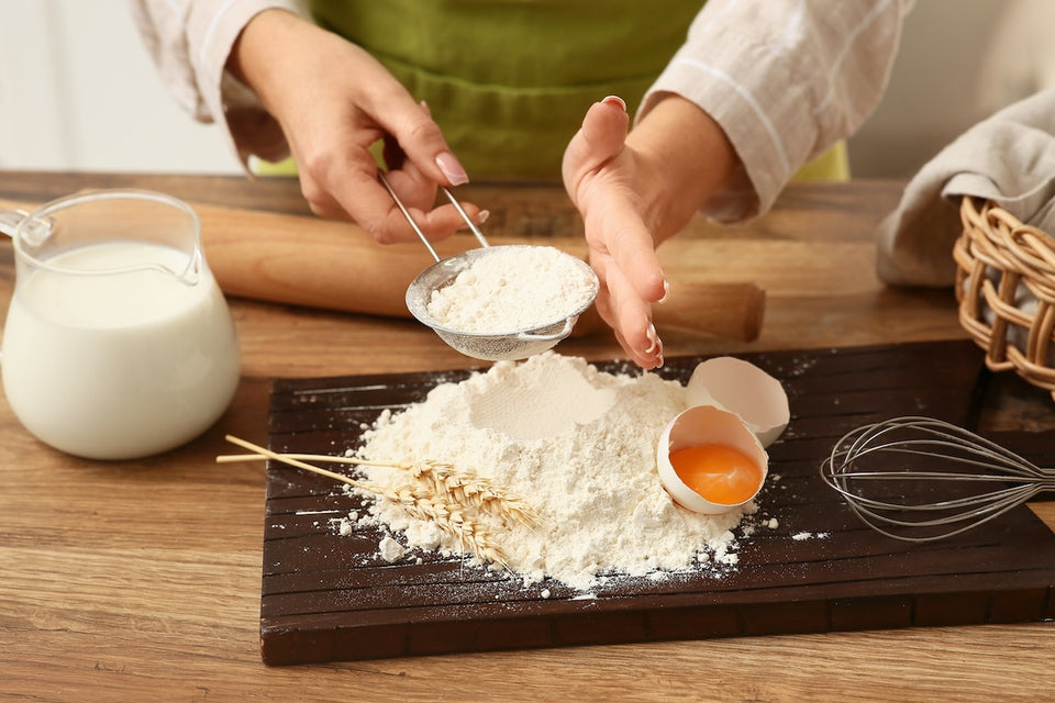 a pair of hands sieving flour with baking ingredients nearby